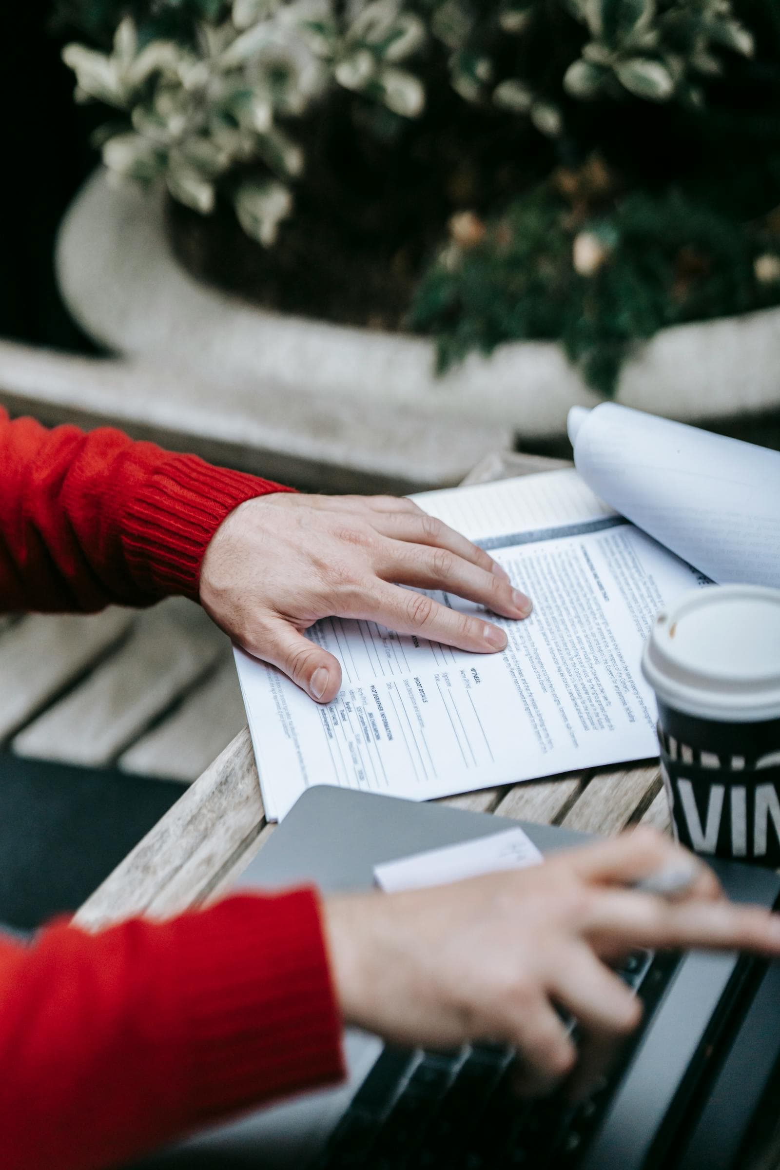 Hands reviewing paperwork beside a laptop and coffee cup on an outdoor table.